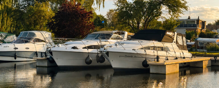 Two white motorboats docked at a marina, secured with ropes. The boats are situated on calm water, with greenery and trees in the background. A building is visible in the distance, adding to the serene marina setting.