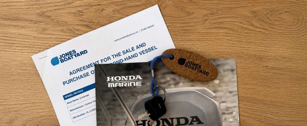 A document titled 'Agreement for the Sale and Purchase of New and Second-Hand Vessel' from Jones Boatyard, along with a Honda Marine brochure and a key. The items are placed on a wooden surface.