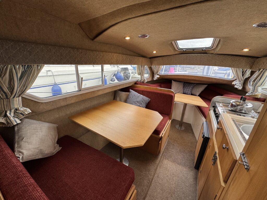 Interior of a small boat featuring a compact dining area with a wooden table and red upholstered benches. Large windows with curtains allow natural light. A small kitchen area with a sink and storage is visible on one side.
