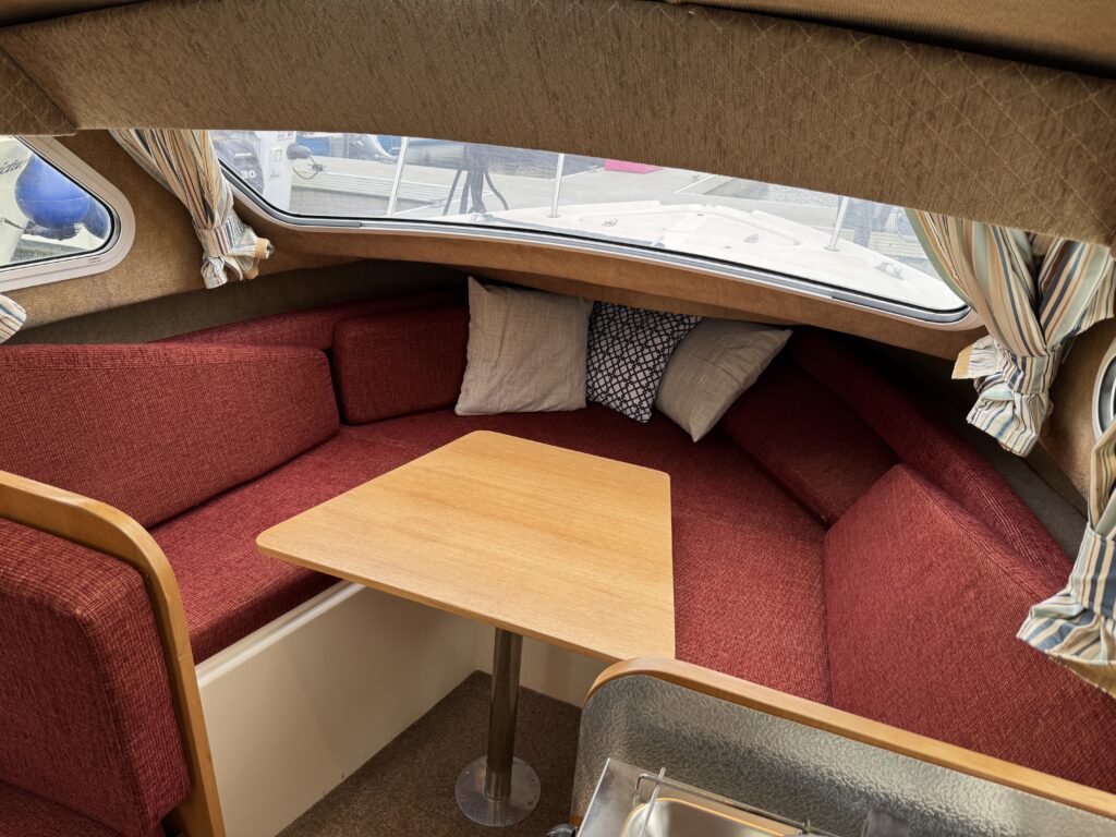 Interior of a small boat featuring a cozy seating area with red fabric cushions arranged around a wooden table. Two throw pillows are placed against the wall under a window with striped curtains, providing natural light.