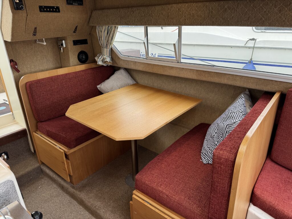 Interior of a boat featuring a dining area with a wooden table surrounded by two red upholstered benches. One bench has a gray and white patterned pillow. A window above allows natural light, with boat features visible outside.