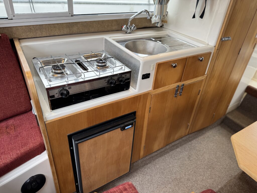 A compact kitchen area of a camper van featuring a stainless steel sink, a two-burner gas stove, and a small refrigerator. The cabinetry is made of wood, and there is a red cushioned seating area visible to the left.