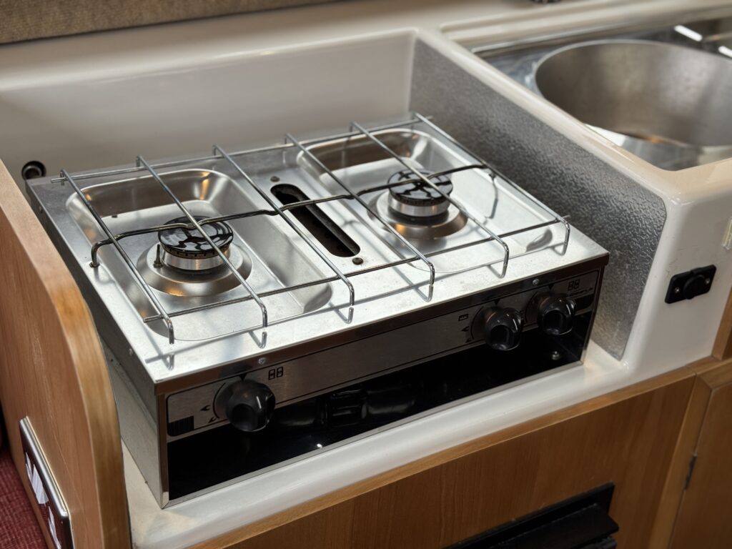 A silver two-burner gas stove installed in a wooden kitchen cabinet. The stove features metal grates above each burner, with control knobs on the front. A stainless steel bowl is visible adjacent to the stove.
