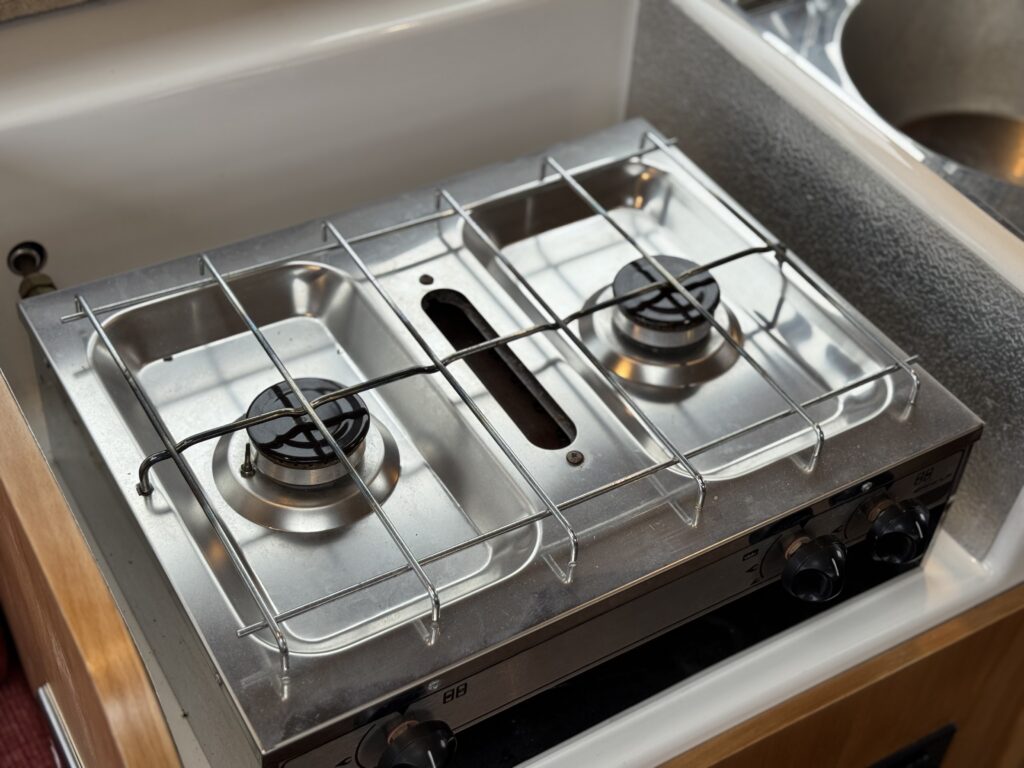 A stainless steel two-burner gas stove integrated into a countertop. The stove features two black control knobs and a protective grating over the burners. The surrounding area includes a portion of a sink in the background.