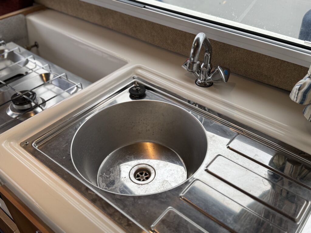 A stainless steel sink with a circular basin is installed in a kitchen countertop. A faucet with a single handle is positioned above the sink. To the left, there are two gas burners visible, and light is coming in through a window above the sink.