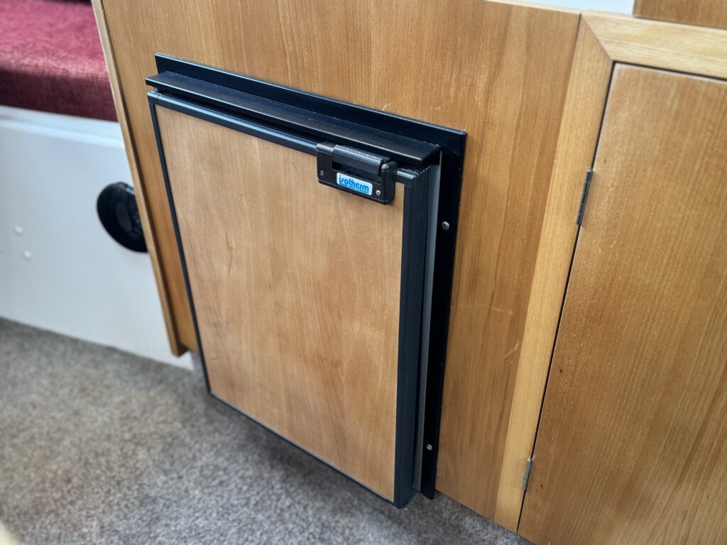 A compact wooden cabinet door with a black handle and metal frame, mounted in a boat interior. The door, slightly ajar, reveals the wooden panel texture. The surrounding area includes a plush red seat and soft carpet.