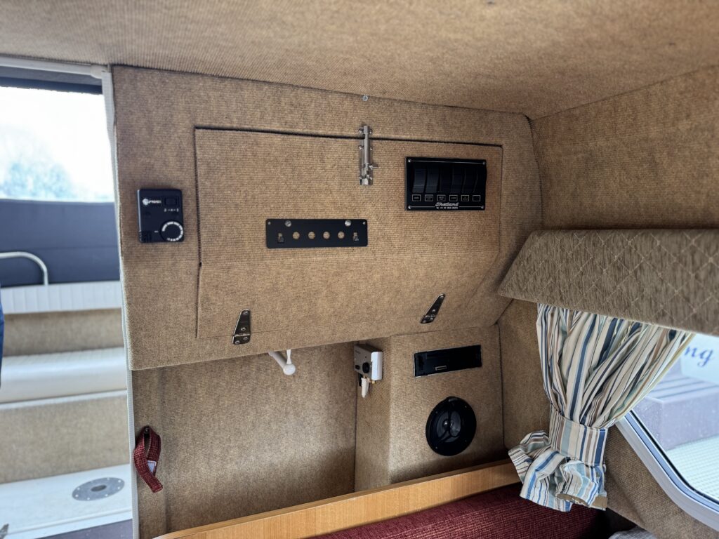 An interior view of a recreational vehicle featuring a textured brown wall panel. Visible components include a control panel with buttons and a digital display, several switches, and a power outlet. A partially opened window with striped curtains is also present.