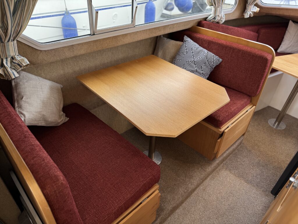 Interior view of a boat with a cozy seating area. Two red cushioned benches are positioned around a hexagonal wooden table. Light-colored walls and large windows provide natural light, with decorative blue objects visible outside.