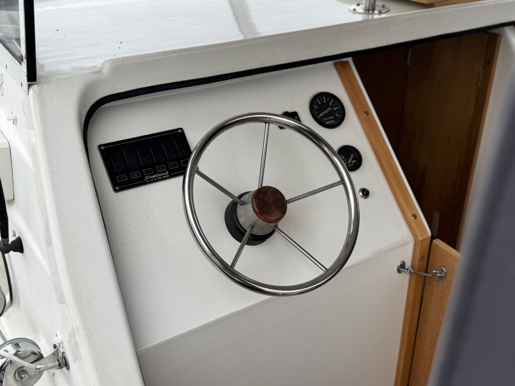 A view of a boat's helm, featuring a stainless steel steering wheel, a control panel with digital display, and various gauges. The dashboard is white with wooden accents, and the setting appears well-maintained, suggesting a focus on navigation and control.