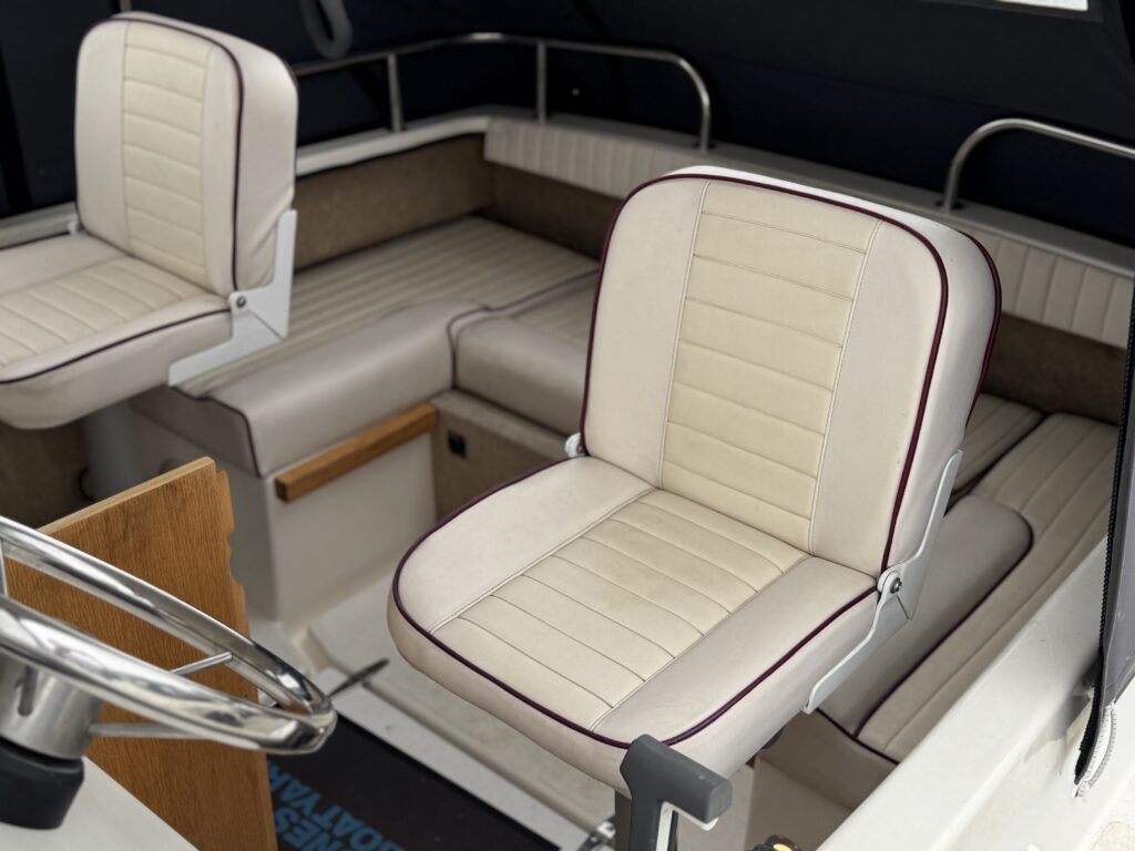 Two upholstered boat seats in light beige with horizontal stitching, located in a cabin area. A steering wheel is visible in the foreground, and the interior features a dark wall and wooden accents.