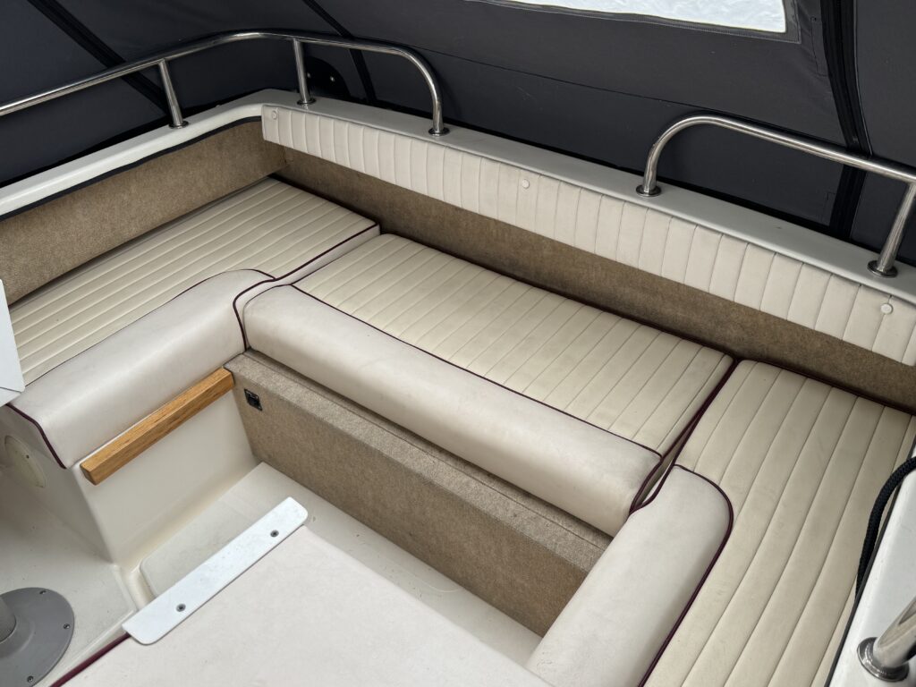 Interior of a boat featuring cushioned seating with beige and brown accents. The cushions have a quilted design and are arranged in a U-shape. There is a wooden panel on one side and a dark canopy visible overhead.