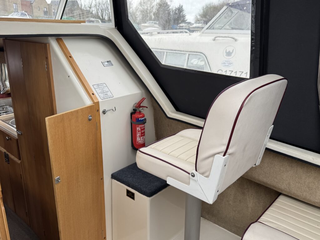 Interior view of a boat with a captain's chair upholstered in white and maroon, positioned near a cabinet. A fire extinguisher is mounted on the wall, and the background shows boats through a window. Bright natural light illuminates the space.
