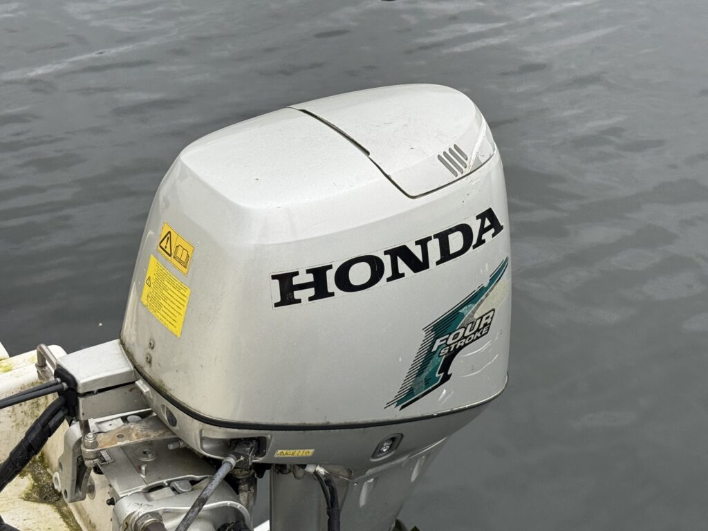 A close-up view of a Honda outboard motor mounted on a boat, showing the motor's metallic surface and branding. The water's surface is visible in the background, reflecting the motor, indicating a marine environment.