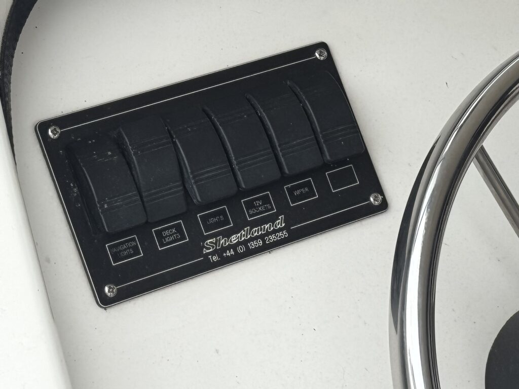 A black control panel with five switch toggles mounted on a white surface, labeled for various functions such as 'Back', 'Lights', 'Main', 'Winch', and 'Emergency'. Next to the panel is a round, silver steering wheel.
