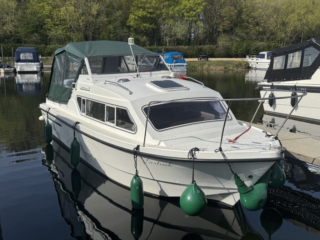 A white cabin cruiser boat is docked at a marina, featuring a green canopy over the wheelhouse. It has green mooring fenders attached and is surrounded by calm water reflecting nearby trees and other boats.
