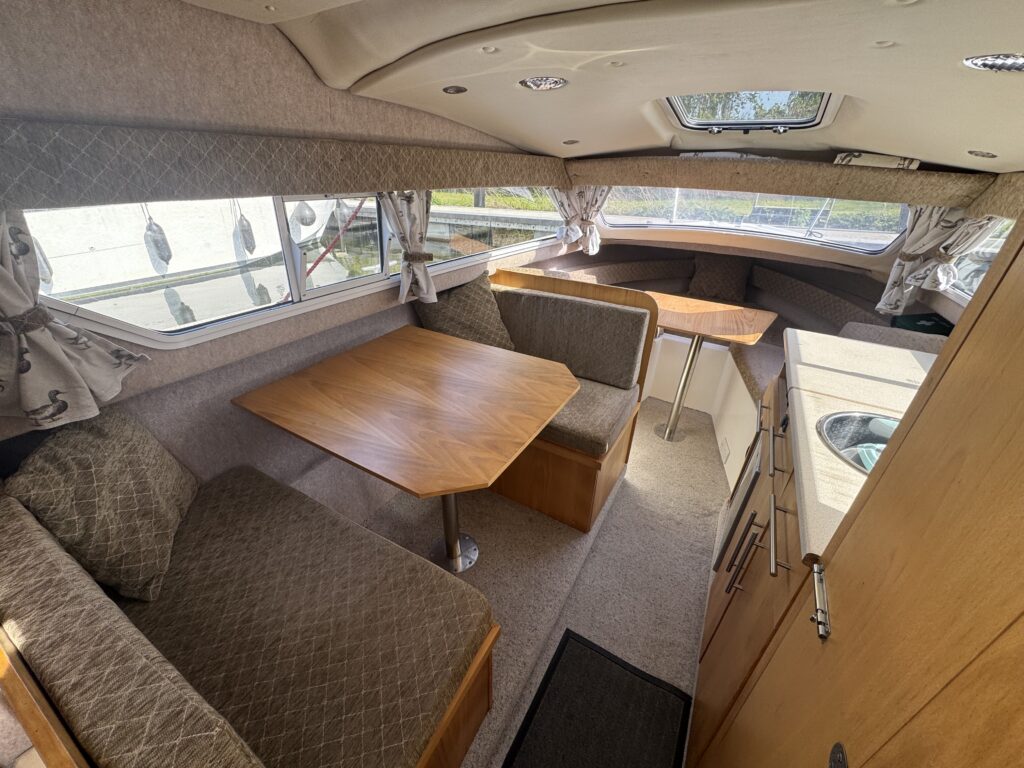 Interior view of a small boat cabin, featuring a U-shaped seating area with a wooden table and grey upholstery. A kitchenette is visible, including a sink, with windows allowing natural light. Curtains frame the windows and there is carpet on the floor.