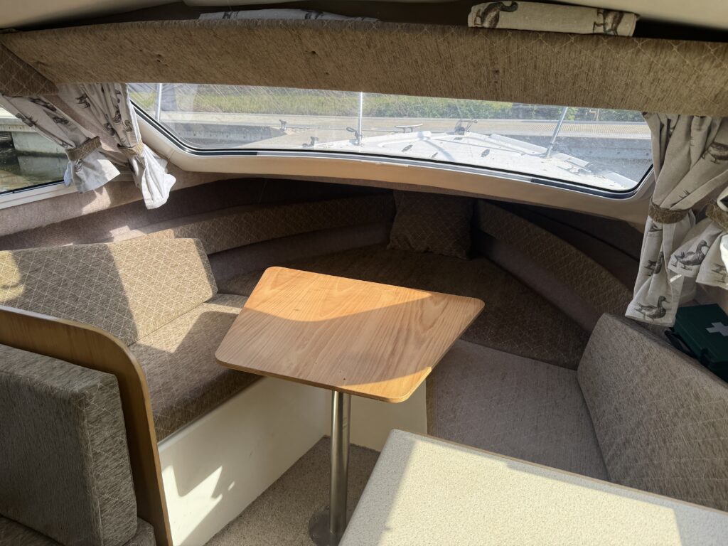 Interior of a boat featuring a U-shaped seating area with light brown upholstery and patterned curtains. A wooden table stands in the center, with windows allowing natural light, and a carpeted floor. A second table is visible in the foreground.
