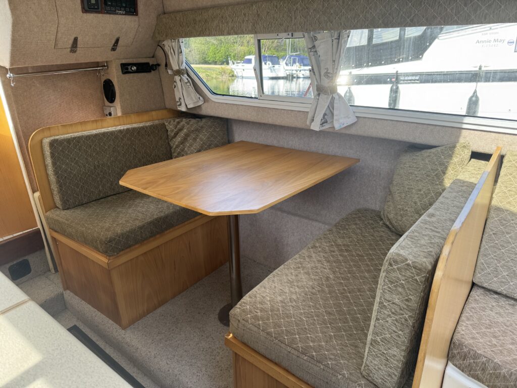 Interior of a boat featuring a small dining area. Two upholstered bench seats are positioned around a wooden table, with sunlight coming through windows and a view of a marina outside. The decor is simple, with pale walls and light-colored upholstery.