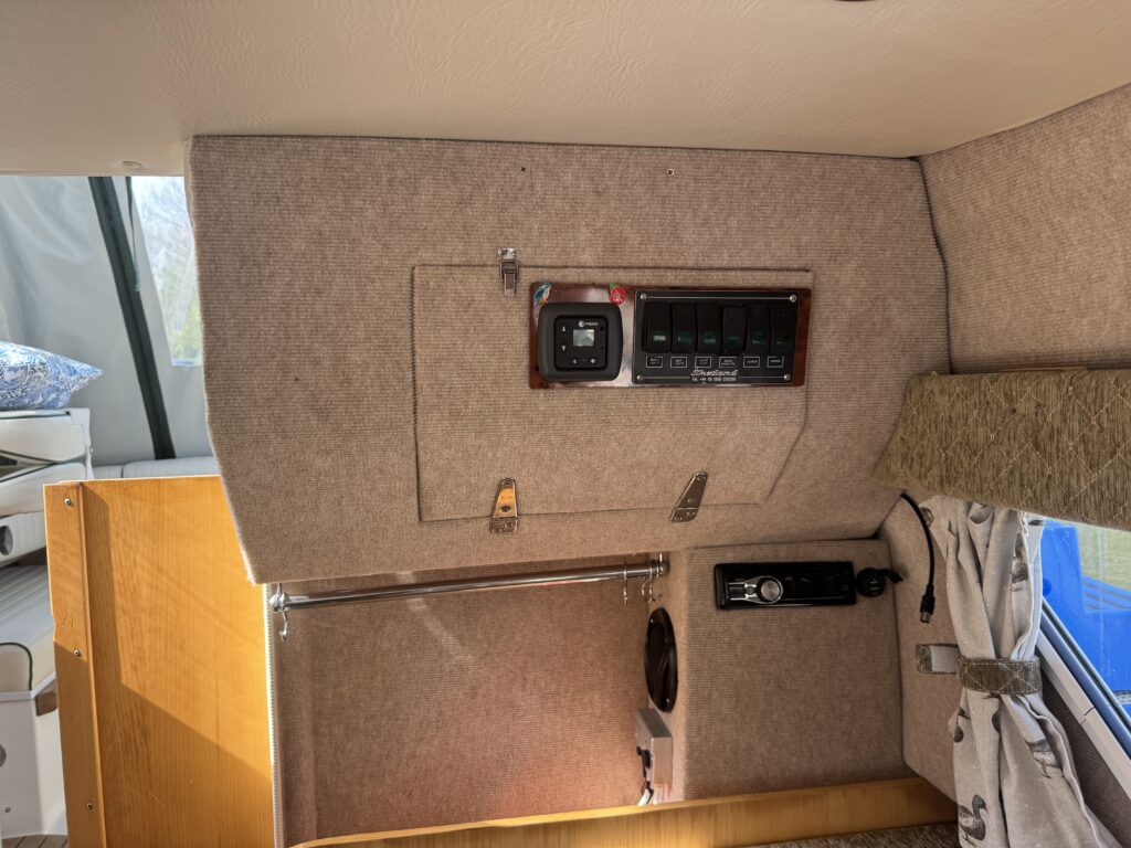 Interior of a camper showing control panel and fixtures. The wall features a switch panel with labeled buttons and a power outlet. Nearby, a metal rod for hanging items is visible, along with a speaker and fabric-covered window.