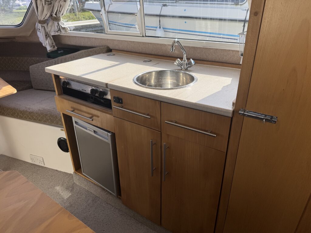 A compact kitchen area in a boat featuring a countertop with a sink, a stove, and a refrigerator built into wooden cabinets. Natural light comes through windows, revealing a seating area in the background and boats visible outside.