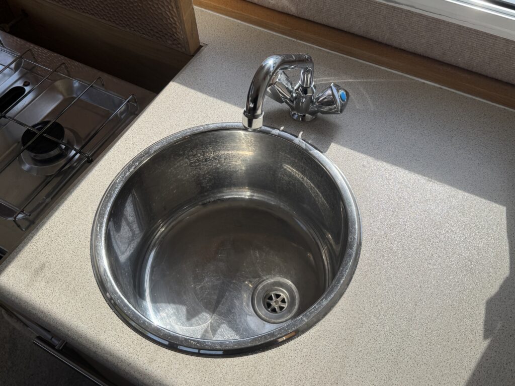 A stainless steel sink with a circular basin is positioned on a countertop. The faucet is seen on the right side above the sink. A gas stove is partially visible to the left, and sunlight illuminates the countertop surface.
