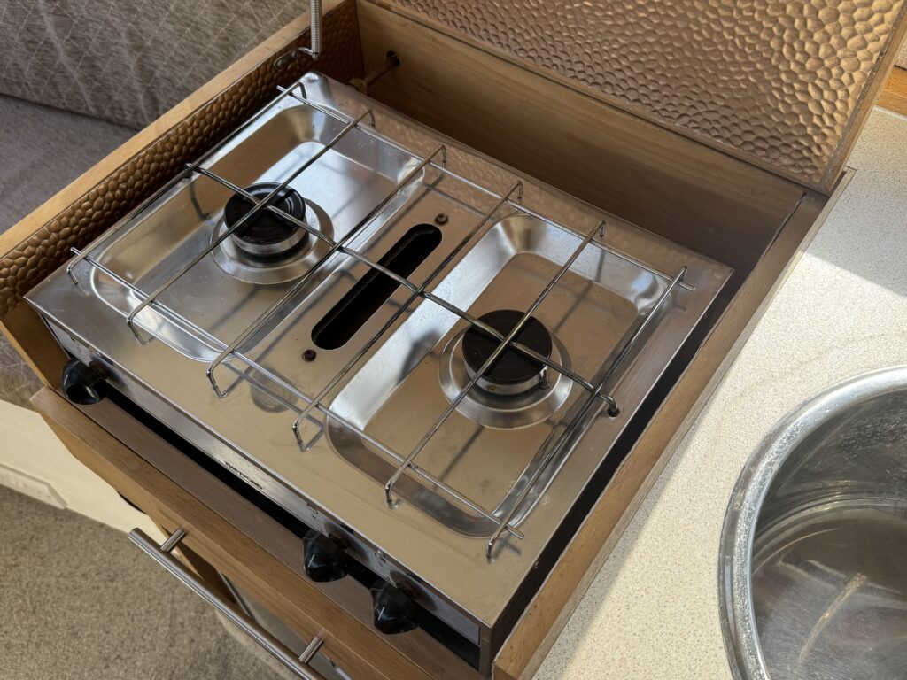 A stainless steel two-burner stove integrated into wooden cabinetry, positioned next to a round sink. The stove features two burners with metal grates and a black control knob below each burner. A textured brown panel is visible above the stove.