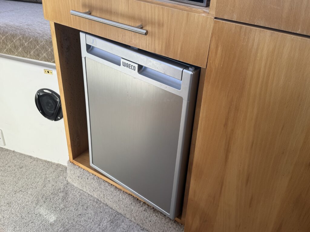 A compact WAECO refrigerator installed in a wooden cabinet with a light wood finish. The refrigerator features a stainless steel front and a simple handle, positioned near a carpeted floor. A sofa can be seen in the background.