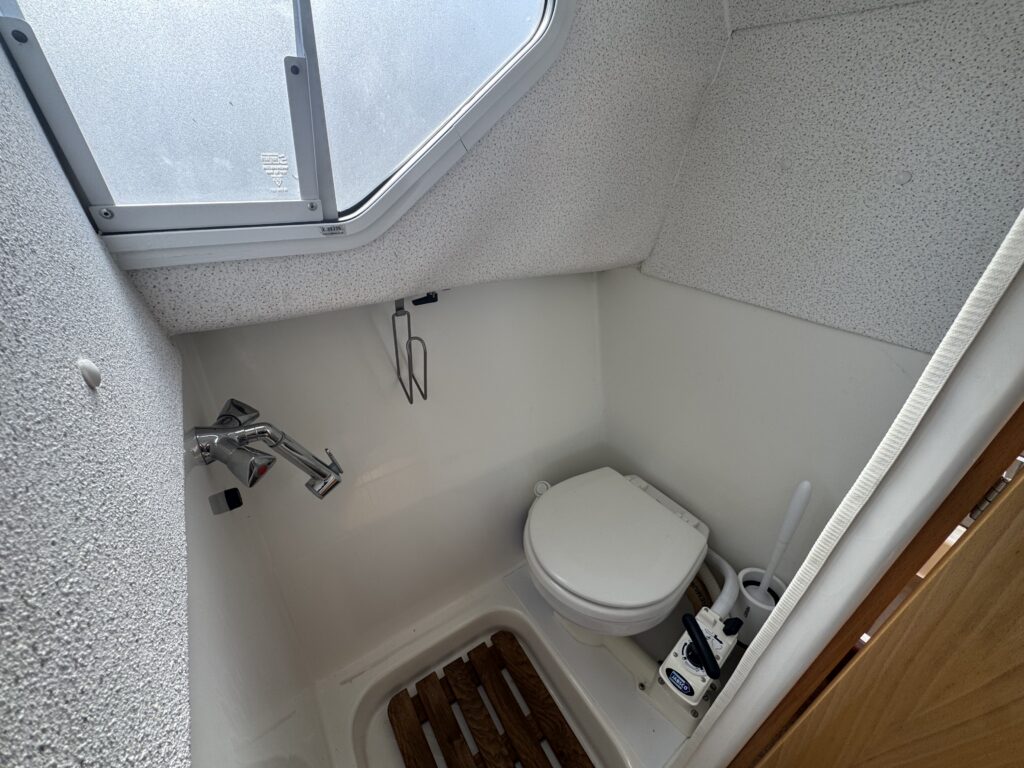 A small bathroom space featuring a toilet, a sink with a faucet, and a window for natural light. The floor has wooden slats, and a toilet brush is visible next to the toilet. The walls are finished with a textured white material.