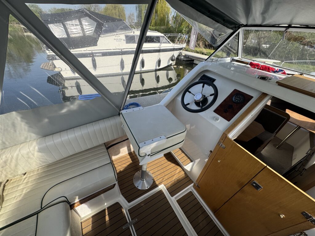 View from the cockpit of a small boat, featuring a steering wheel, a table, and polished wooden surfaces. In the background, another boat is docked on calm water surrounded by trees and a clear blue sky.