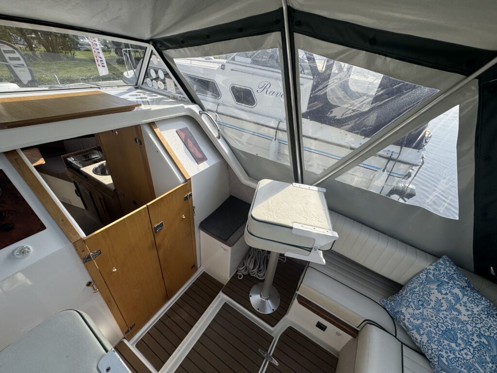 A view from above of an interior cabin of a boat, featuring wooden cabinets, a white table on a metal stand, and cushioned seating with a blue throw pillow. Large windows allow natural light to enter, and another boat is visible outside.