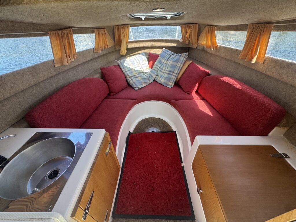 Interior of a boat cabin featuring a red circular sofa with cushions, a small kitchen area with a sink, and a red carpet. Natural light comes through the windows, and the background shows water outside, creating a relaxed boating environment.