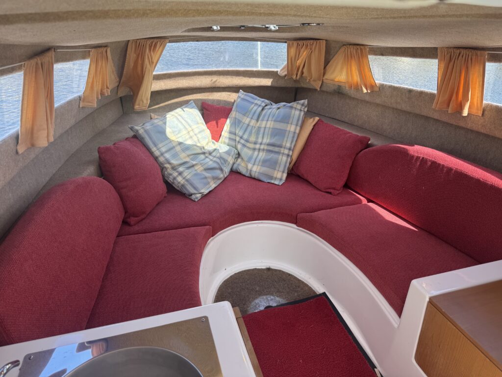 Interior of a small boat with a cozy seating area. The space features red upholstered cushions and several plaid throw pillows. There are curtains along the windows, and a round table is partially visible in the foreground.