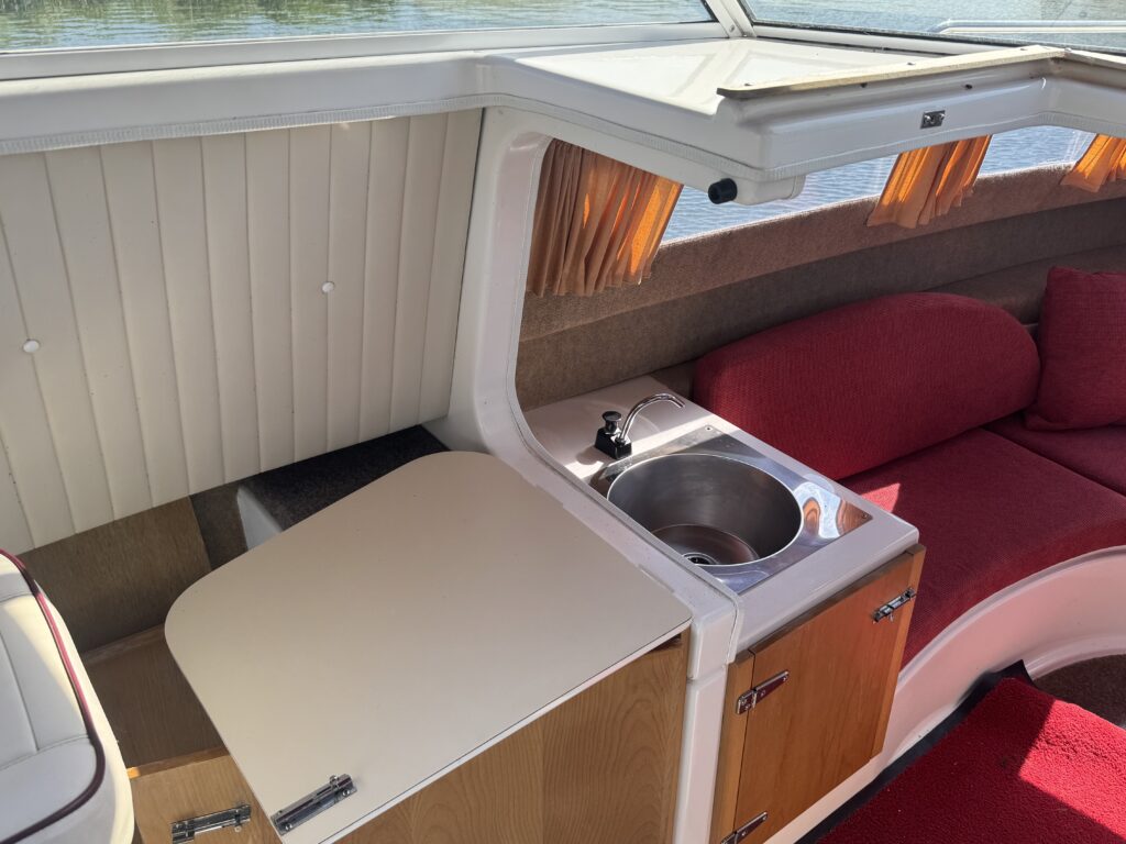Interior view of a boat showing a compact kitchenette area with a circular sink and a white countertop. There are wooden cabinets below, a red cushioned seating area next to the kitchenette, and windows providing natural light.