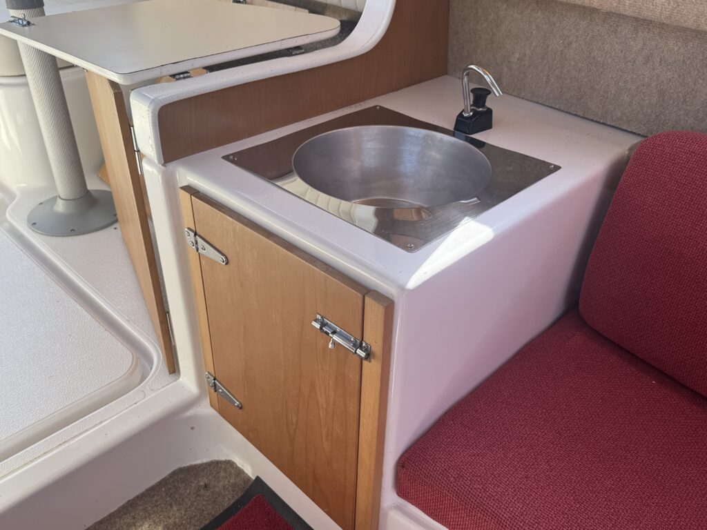 A small kitchenette area on a boat featuring a circular stainless steel sink set into a countertop. Below the sink, there is a wooden cabinet with a single door, and a red cushioned seat is positioned nearby. A faucet is visible.