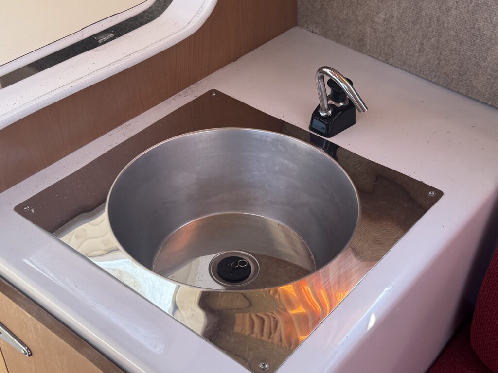 A stainless steel sink with a circular basin and a drain, installed in a wooden cabinet. The faucet is a modern black handle style, placed to the right of the sink. The surrounding surface is smooth and features a light-colored laminate.