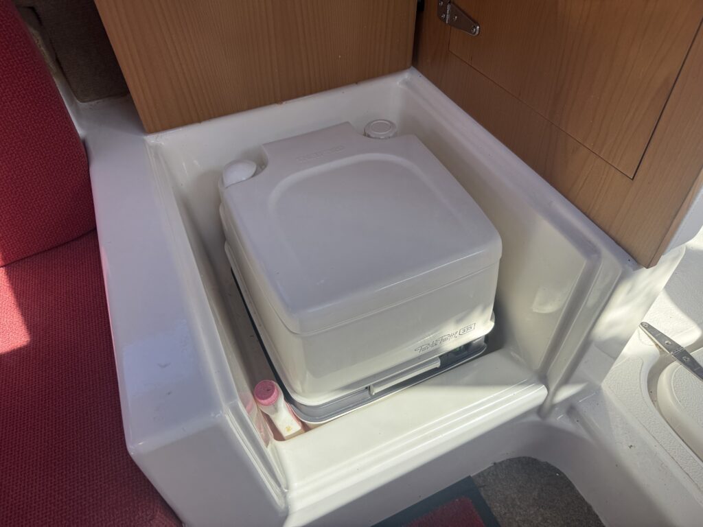 A portable toilet placed in a white base inside a cabin, with red fabric on the adjacent seating area. The cabin wall features wooden paneling, and there is a small pink item near the base of the toilet.