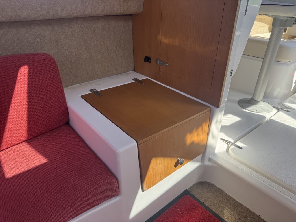 An interior view of a boat featuring a red upholstered seat and a wooden table that can be folded down. The wall behind is beige upholstered, and there is a wooden panel with a latch. Light enters through a nearby window, highlighting the space.