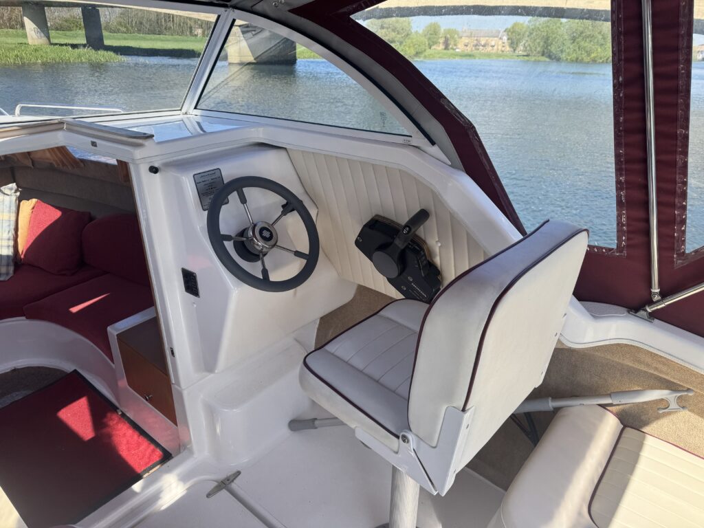 The interior of a boat showing the helm with a steering wheel and throttle control. A white cushioned pilot seat is positioned next to the helm. Large windows provide a view of a river and green landscape outside.