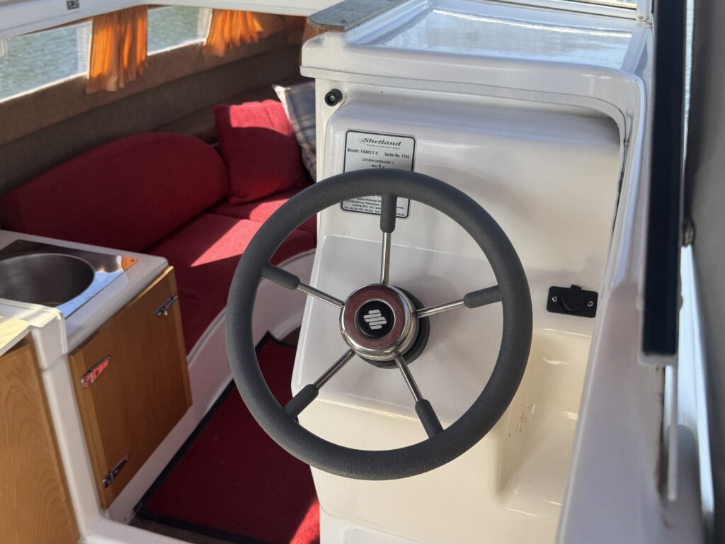 The interior of a boat featuring a steering wheel in the foreground. The helm is white, with a control panel nearby. A cushioned seating area is visible in the background, adorned with red cushions and a wooden cabinet is to the left. Natural light is entering through a window.