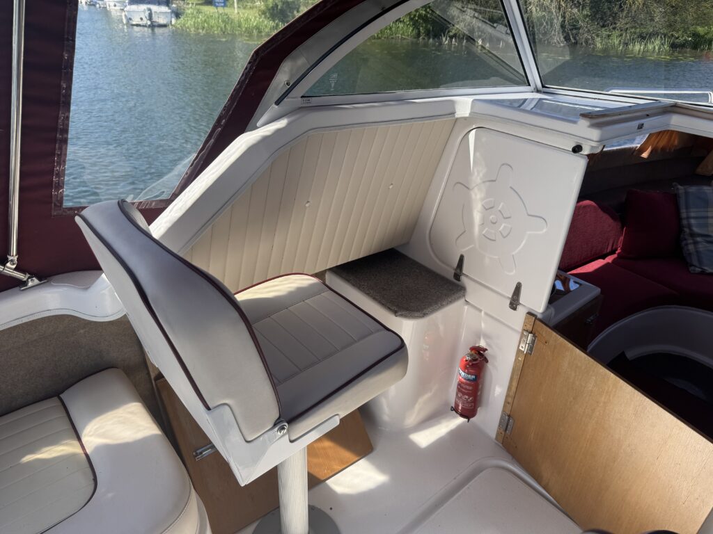 Interior view of a boat's cockpit featuring a single cushioned seat with a tall back, positioned next to a wooden panel. The cockpit has natural light coming in, with a view of water and greenery through large windows.