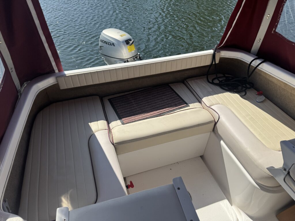 Interior view of a boat seating area with light-colored, cushioned benches arranged around a central space. In the background, a Honda outboard motor is visible on the water, with a red canopy providing shade.