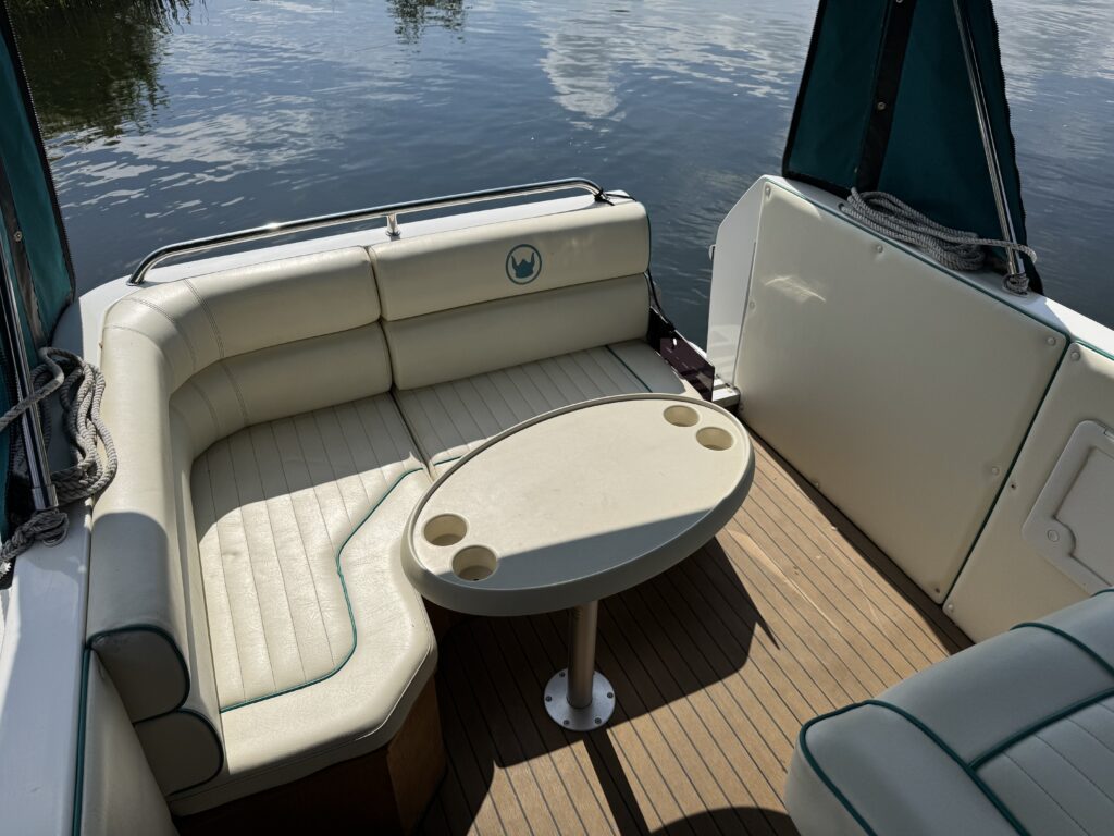 An interior view of a boat's seating area featuring a white cushioned sofa with green trim and a circular table with cup holders. The area is well-lit by sunlight, with water visible in the background.