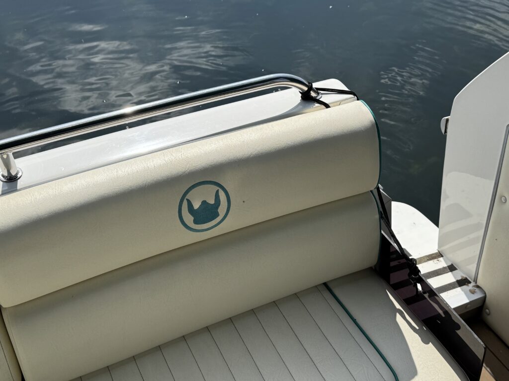 Close-up of a boat's cushioned seat with a circular logo featuring a crown. The seat is beige with green trim, and part of a railing is visible. The water's surface reflects light, creating a serene background.