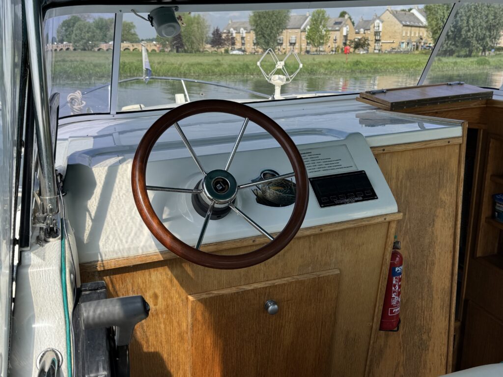 Interior view of a boat's steering area, featuring a wooden steering wheel, control panel, and upholstered seating. A fire extinguisher is mounted nearby. In the background, there's a calm water scene with buildings lining the shore.