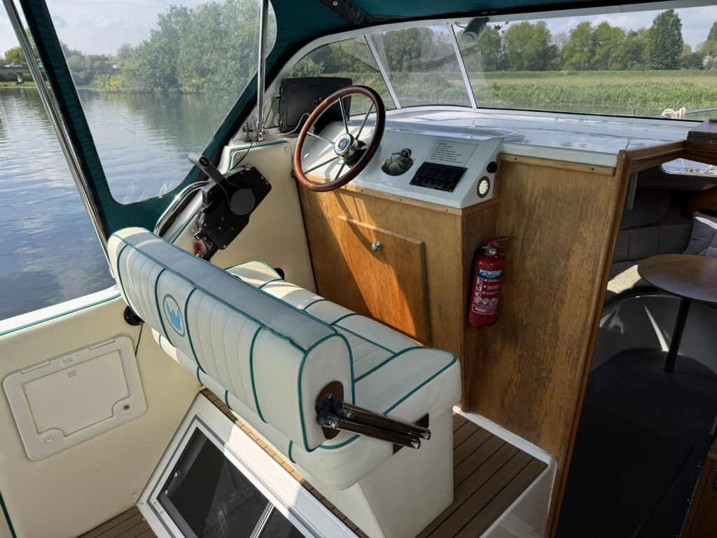 Interior of a boat with a view of water outside. The helm includes a steering wheel and a control panel. A padded seat with blue and white stripes is positioned at the helm, and there is a wooden cabinet nearby. A fire extinguisher is mounted on the wall.