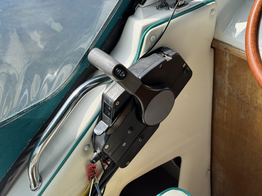 A close-up view of a boat's throttle control panel, featuring a black throttle lever and several buttons. The panel is mounted on a light-colored surface, with visible wiring and a wooden steering wheel in the background. Natural light reflects on the surface.