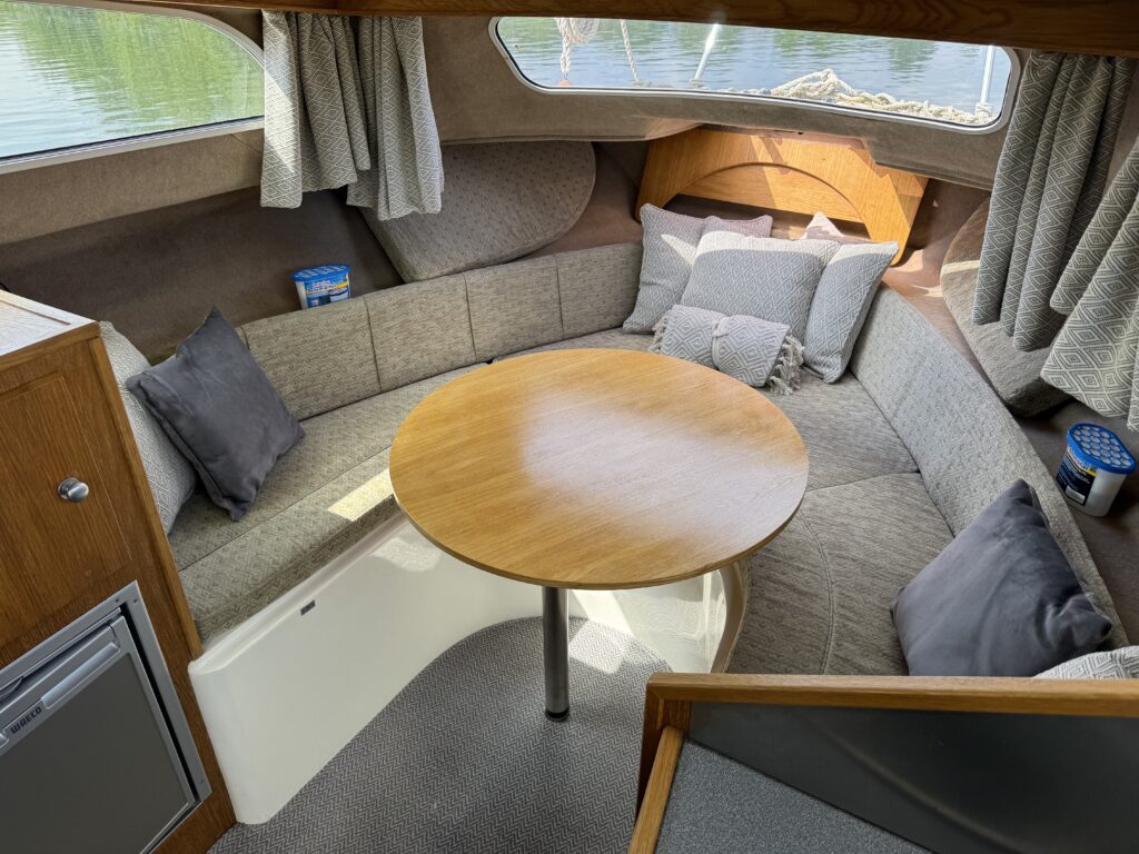 An indoor seating area of a small boat, featuring a round wooden table surrounded by cushioned benches. The space is decorated with gray throw pillows and has curtains partially drawn. A view of water is visible through a window.