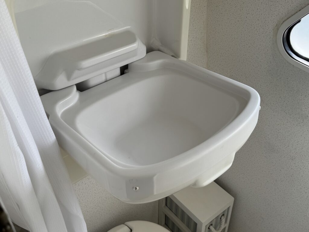 A white sink with a smooth surface, built into a wall in a small bathroom. The sink is positioned above a toilet and a storage unit can be seen nearby. Natural light is entering through a small window, illuminating the area.