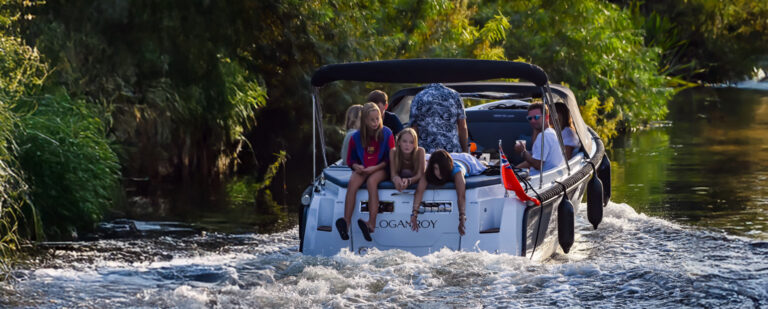 A group of five people relaxing on a boat as it moves along a calm river, surrounded by lush greenery. Two adults are seated in the front, while three children sit on the back, enjoying the ride.