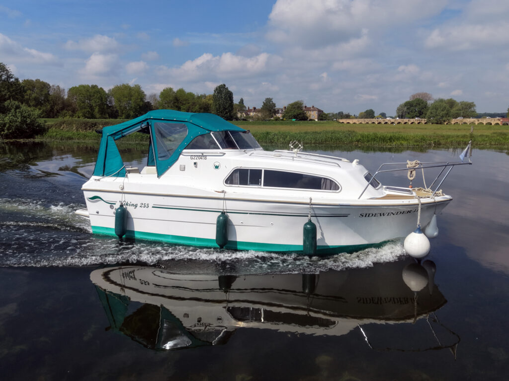 A white motorboat with a teal canopy is cruising on a calm river. The boat features two fenders and a buoy. Lush greenery and trees are visible along the shore, with a partly cloudy sky reflected in the water.